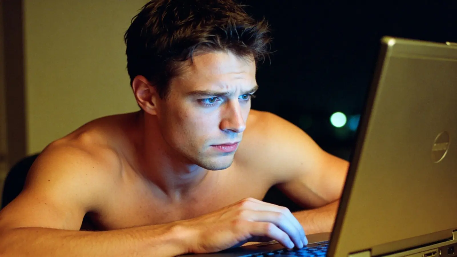 Young man at night lit by a laptop glow with a conflicted expression